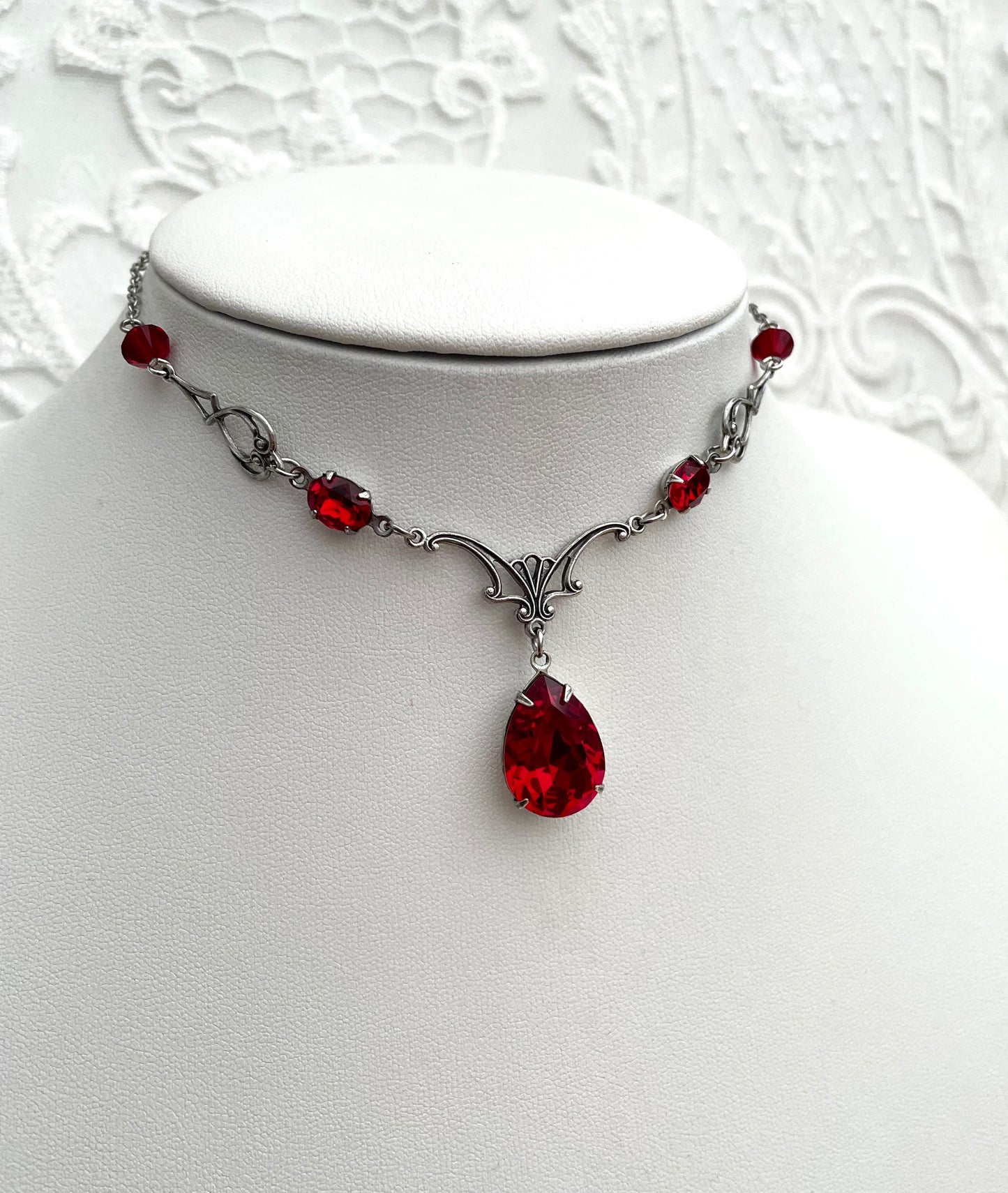 A silver necklace with a large teardrop-shaped red gemstone pendant, smaller red accent gemstones along the chain, and intricate silver detailing.