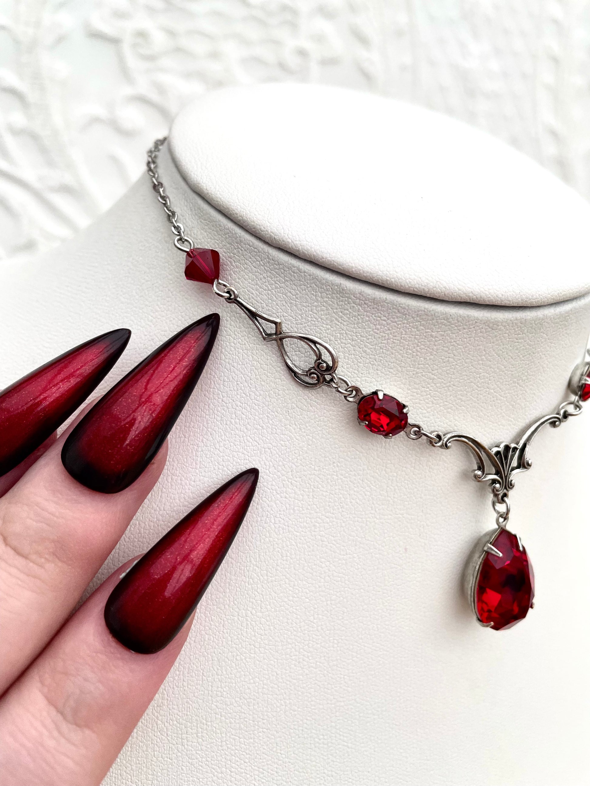 A silver necklace with a large teardrop-shaped red gemstone pendant, smaller red accent gemstones along the chain, and intricate silver detailing.