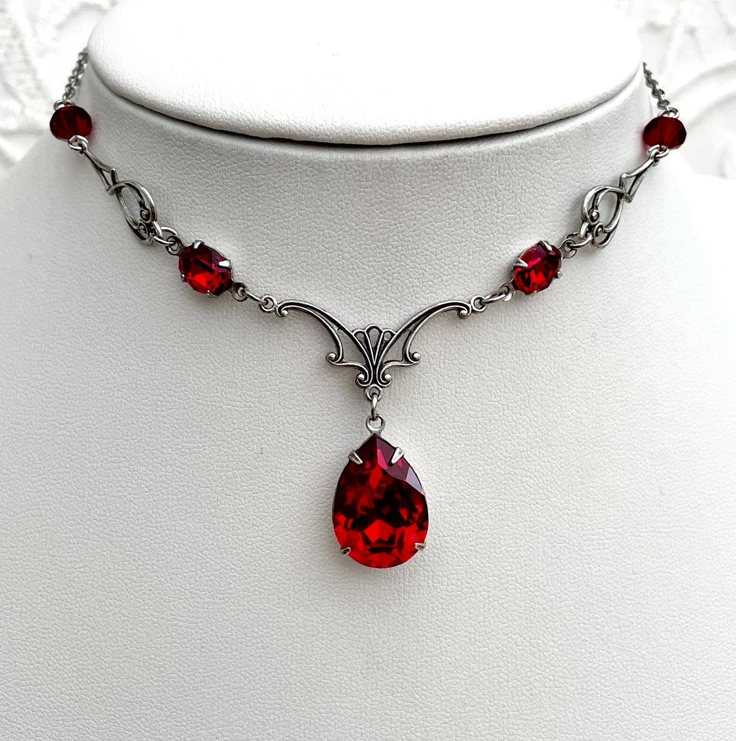 A silver necklace with a large teardrop-shaped red gemstone pendant, smaller red accent gemstones along the chain, and intricate silver detailing.