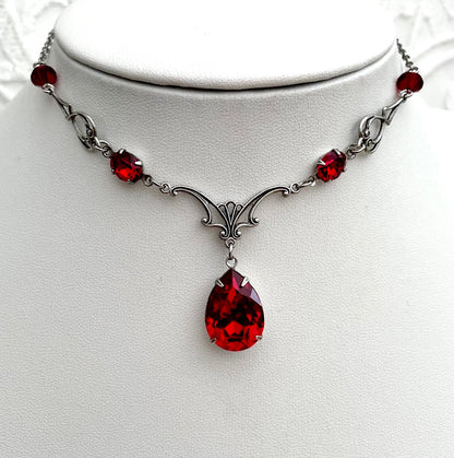 A silver necklace with a large teardrop-shaped red gemstone pendant, smaller red accent gemstones along the chain, and intricate silver detailing.