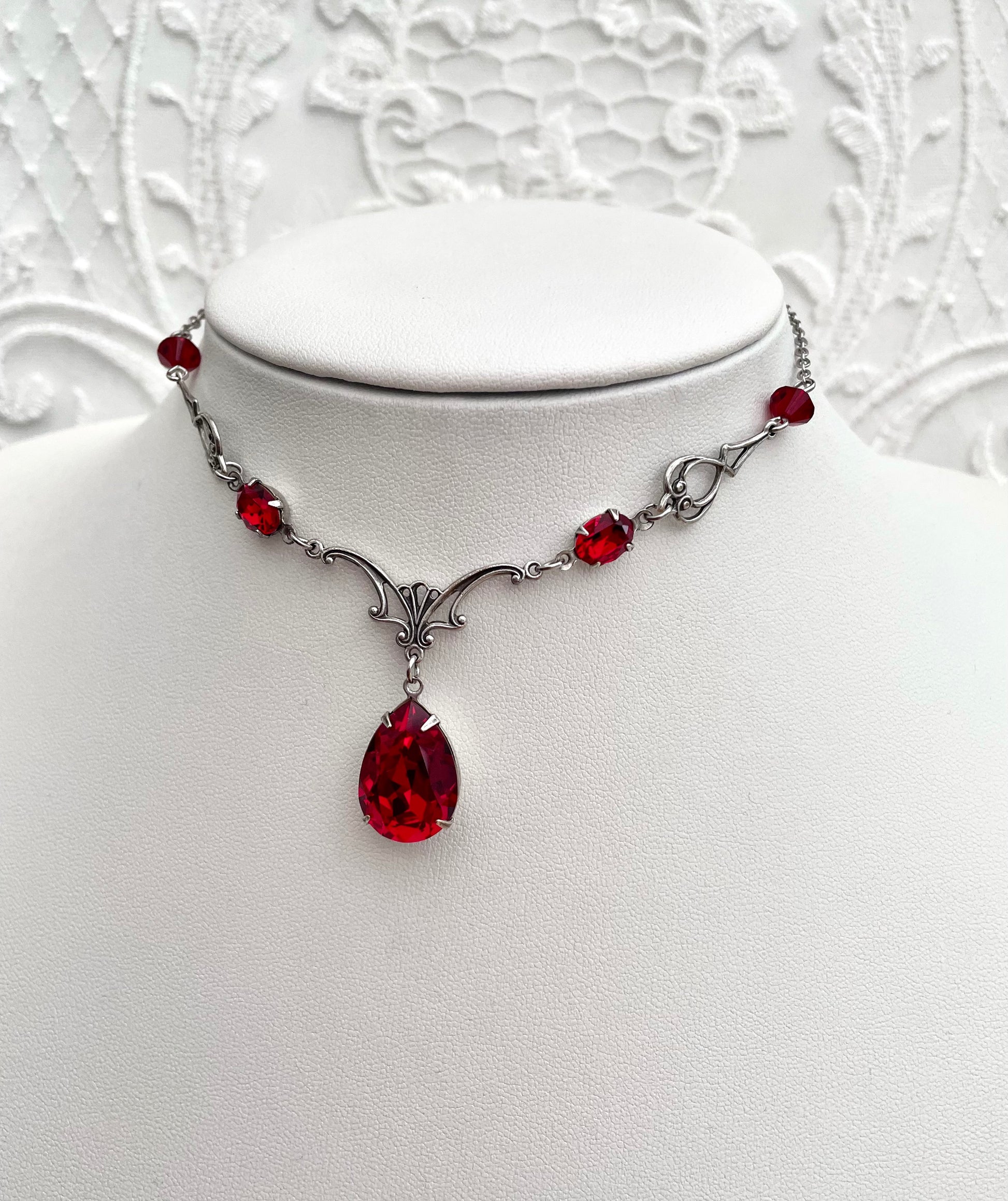 A silver necklace with a large teardrop-shaped red gemstone pendant, smaller red accent gemstones along the chain, and intricate silver detailing.