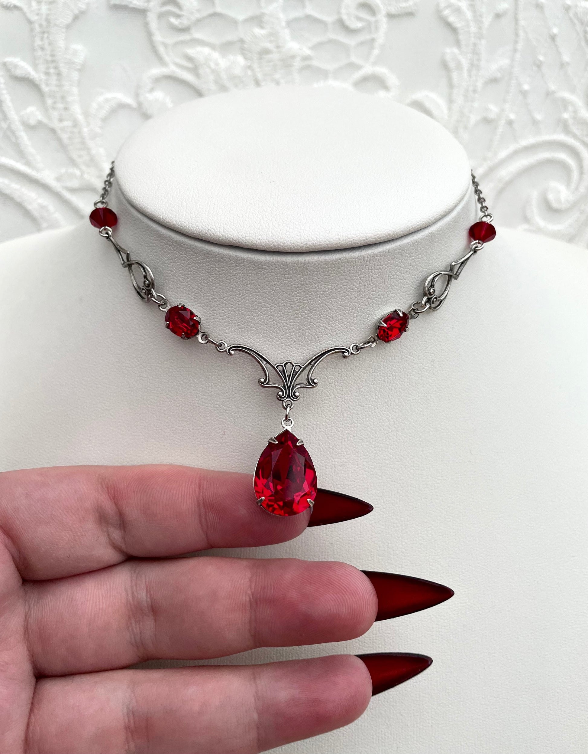 A silver necklace with a large teardrop-shaped red gemstone pendant, smaller red accent gemstones along the chain, and intricate silver detailing.