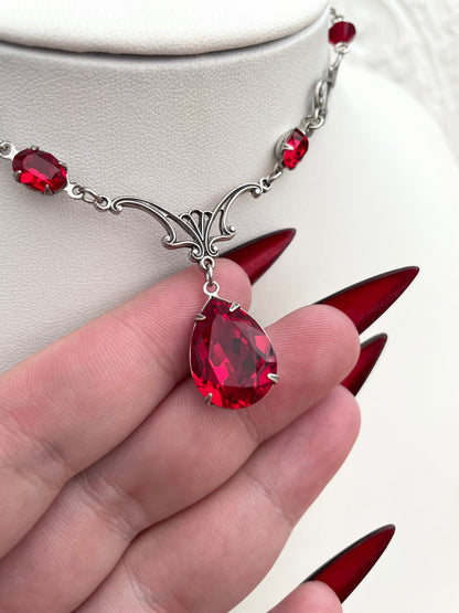 A silver necklace with a large teardrop-shaped red gemstone pendant, smaller red accent gemstones along the chain, and intricate silver detailing.